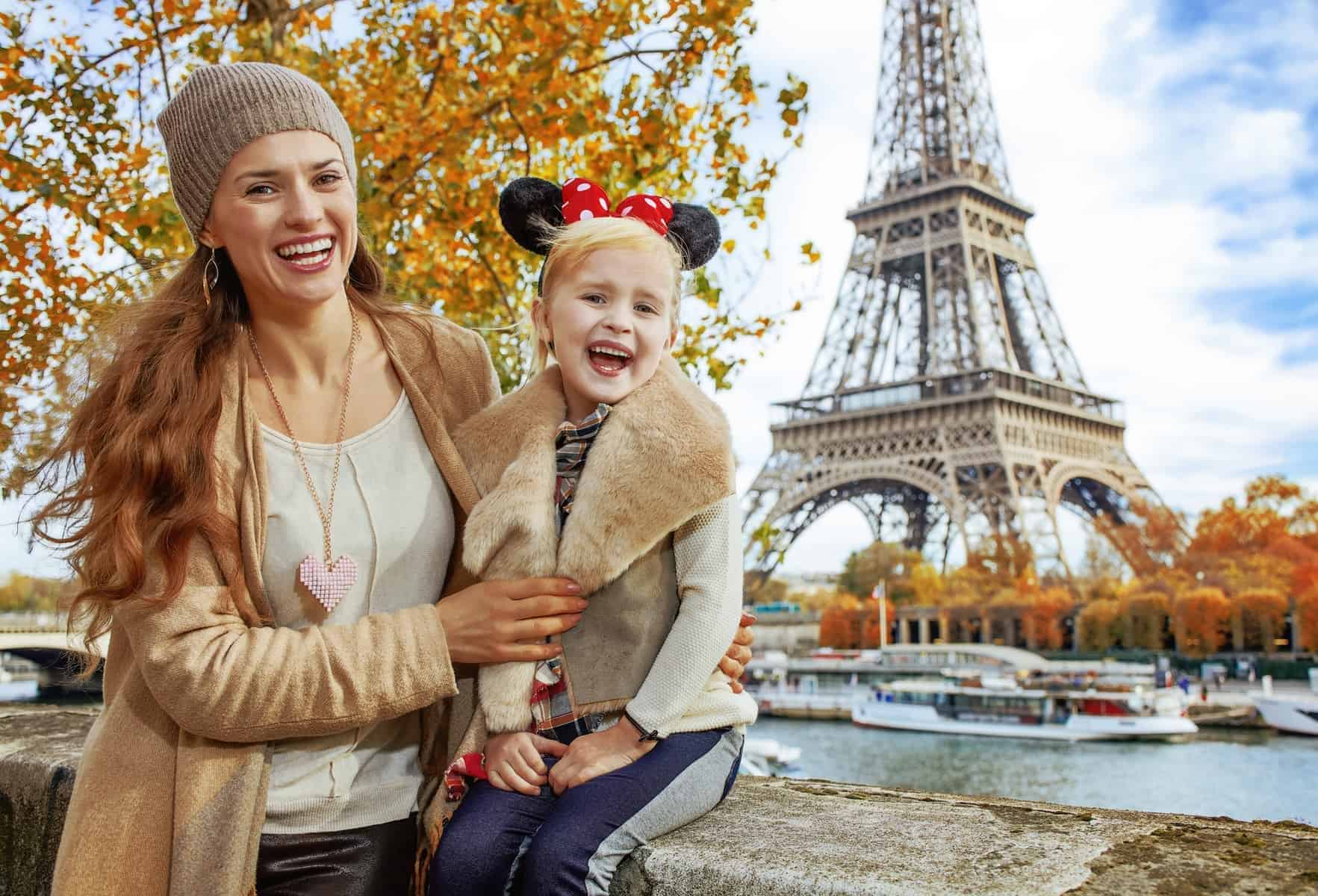 Perfect autumn holidays in Disneyland and Paris. Portrait of smiling tourists mother and daughter in Minnie Mouse Ears on embankment near Eiffel tower in Paris, France sitting on the parapet
