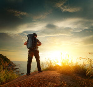 Book din rejse. traveltalk.dk. Hiker with backpack standing on a rock and enjoying sunset over sea